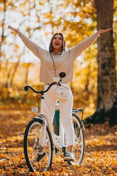 Young woman in cozy outfit rejoices autumn, freedom and beautiful nature. Girl with open hands. Love, gratitude concept. Amazing scene of trendy girl with bicycle. High quality photo