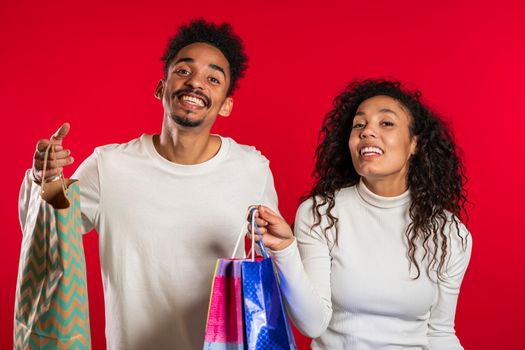 Happy young african couple with colorful paper bags after shopping isolated on red studio background. Seasonal sale, purchases, spending money on gifts concept.