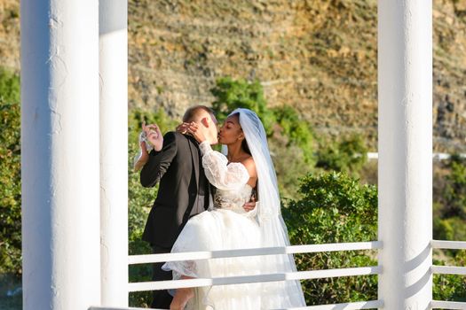 Young beautiful interracial newlyweds are kissing in the gazebo, against the backdrop of sunny mountains