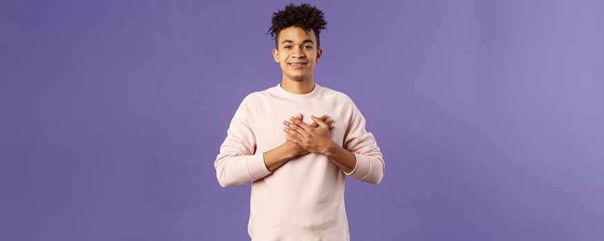 Portrait of romantic handsome hispanic man with dreads, hold hands pressed to heart and smiling pleased, see heartwarming nice gesture, receive lovely gift, being touched, purple background.