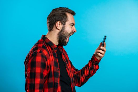 Angry modern hipster in red plaid shirt screaming down his mobile phone. Stressed and depressed man on blue background.