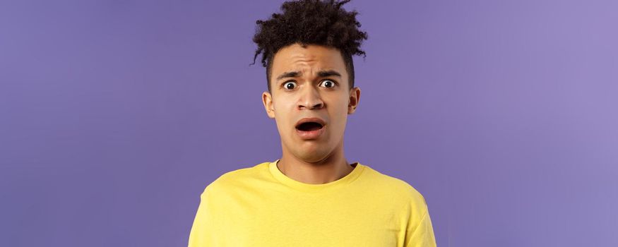 Close-up portrait of shocked, alarmed young man gasping, open mouth scared and frightened, staring camera concerned, facing troublesome shocking news, purple background. Copy space