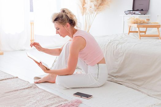 A slender woman, a pink top and white pants, sits on the floor in the morning, near a white bed, working on a digital tablet, a smartphone lies nearby