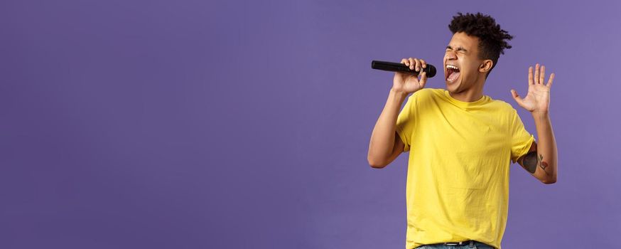 Portrait of passionate carefree young hispanic singer with dreads and tattoos, reaching highest note in song, raising hand up singing loud at microphone with closed eyes, purple background.