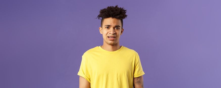 Waist-up portrait of young concerned hispanic man with worried gaze looking at camera, frowning being scared for friend stuck in troublesome situation, want help, stand purple background.