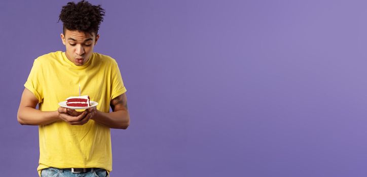 Portrait of cute excited young guy with dreads and tattoos, making wish on birthday, celebrating party, blowing candle on b-day cake, standing purple background dreaming. Holidays concept