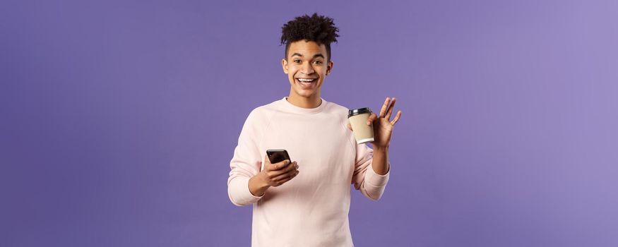 Portrait of cheerful young male student with cup of coffee and mobile phone, waving hand to say hi, smiling friendly as greeting person, meet classmate near university campus, purple background.