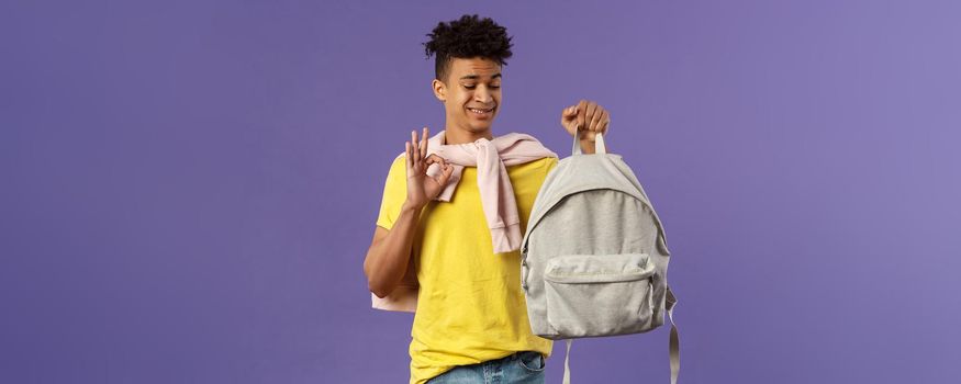 Portrait of young awkward guy, student receive gift new backpack from mom, being displeased with it grimacing but say okay, not bad, looking at rucksack standing purple background.