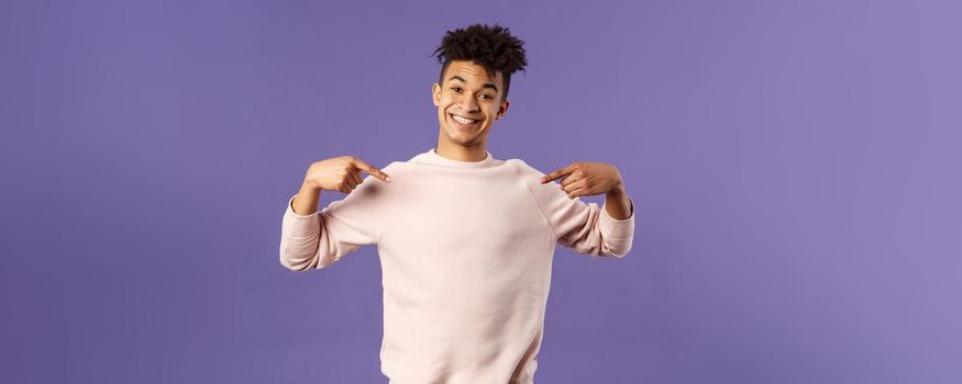 Cute and silly handsome male student pointing at himself and making lovely smile, ready to help offer himself, want participate or being chosen, volunteer to be a candidate, purple background.