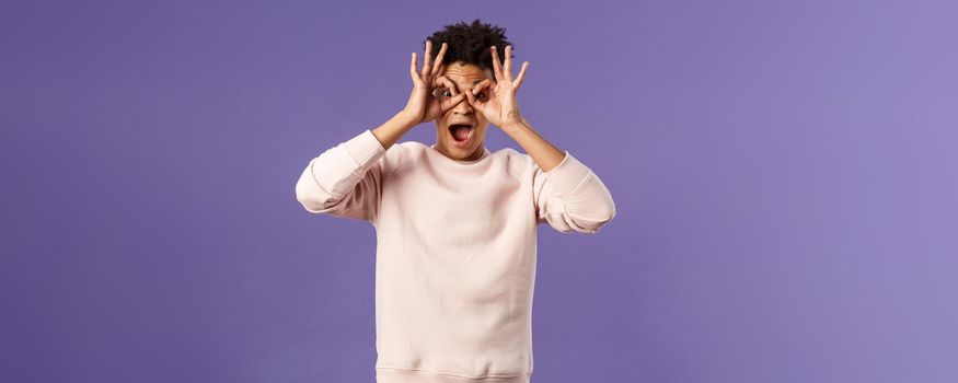 Portrait of funny, enthusiastic young hispanic man fooling around, making finger glasses, mask with hands over eyes, open mouth fascinated and surprised, seeing great product or promo action.