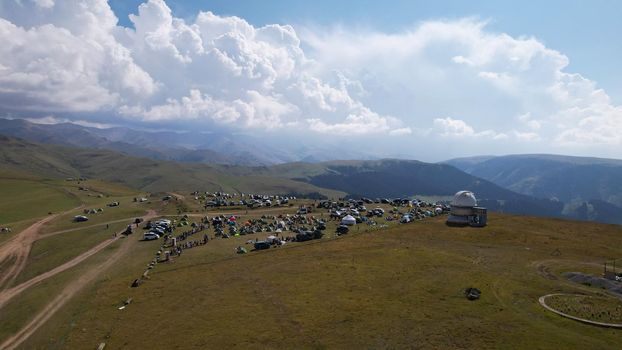 The Assy-Turgen Observatory is high in the mountains. There is a tent camp next to the observatory. Large cumulus clouds in a blue sky. Yellow-green hills, forest in places. Top view from a drone