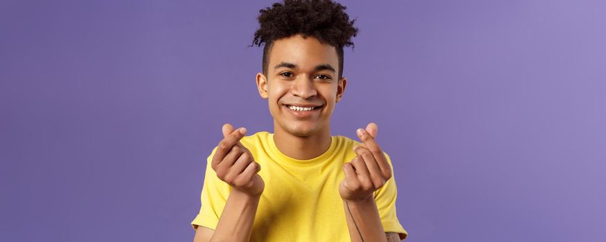 Close-up portrait of lovely cute young man with afro haircut, showing korean heart finger signs and smiling, express his love and sympathy towards person, stand purple background.