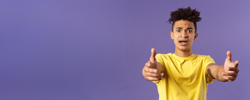 Waist-up portrait of concerned, nervous young man reaching hands forward to hold something but being scared, friend asking take baby for sec, standing very anxious and afraid, purple background.