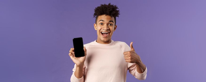 Happy young man, hispanic guy with dreads recommend app, online delivery service or food order, show thumbs-up, smiling excited, holding mobile phone, show smartphone screen.