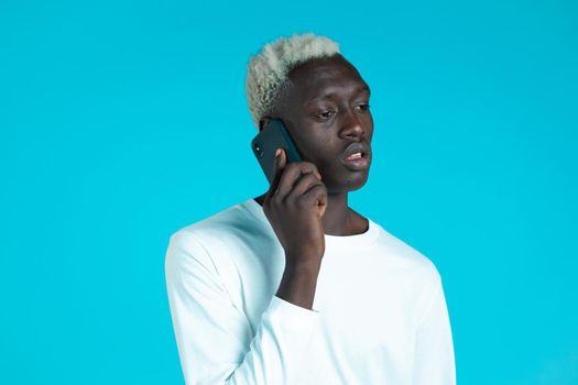 Young afro man speaks serious on phone. Guy holding and using smart phone. Blue studio background
