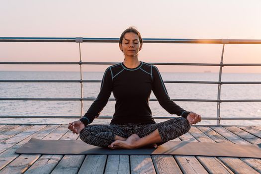 Young brunette woman doing yoga exercises on wooden sea embankment in the morning. Girl in black sports costume. Stretching, practice, healthy lifestyle concept.