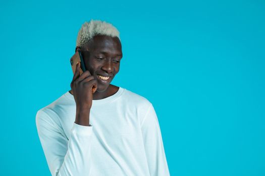 Young african american man speaks with smile on phone. Guy holding and using smart phone. Blue studio background