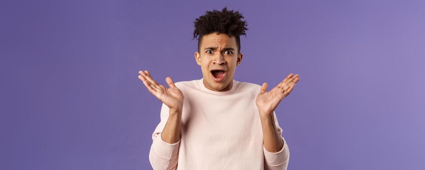 Close-up portrait of displeased, bothered frustrated hispanic man spread hands sideways in dismay and confusion, staring camera upset with disappointed grimacing expression, purple background.