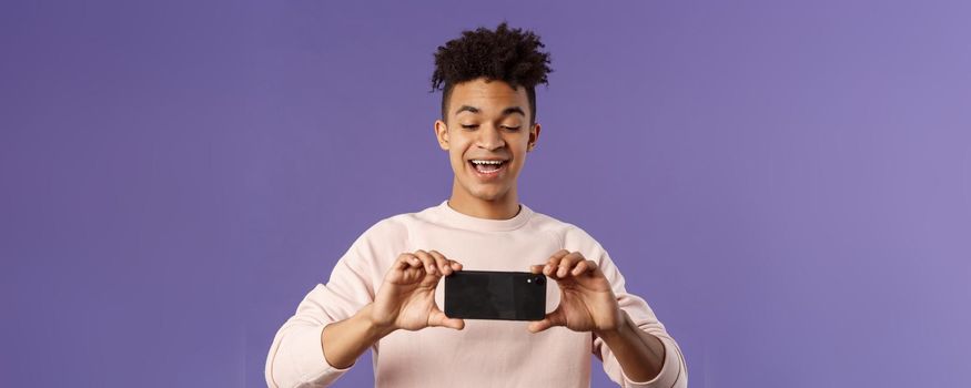 Portrait of amazed, excited young man seeing something interesting, stream concert to his internet social network profile, taking photo or recording video with mobile phone, purple background.