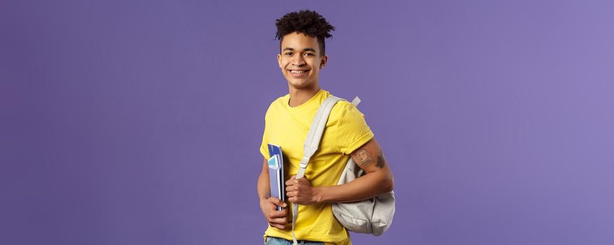 Back to school, university concept. Portrait of handsome charismatic hispanic guy, student walking to campus, going for lecture, studying hold backpack and notebooks, stand purple background.