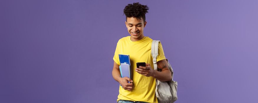 Back to school, university concept. Portrait of pleased good-looking male student texting friend, looking at mobile phone on his way to classes in campus, hold notebooks and backpack.