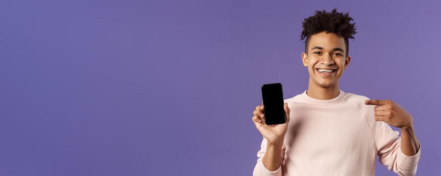 Close-up portrait of young handsome man promoting smartphone app or shopping online, internet delivery for goods, holding mobile phone pointing at display and smiling pleased, recommend.