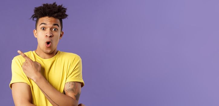 Portrait of surprised, excited young man showing cool new thing, pointing upper left corner, gasping, folding lips, stare camera impressed say wow, standing purple background.