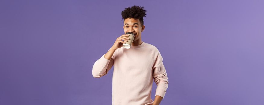 Leisure, lifestyle and people concept. Portrait of young cheerful guy drinking coffee take-away from favorite cafe, smiling enjoying nice spring day, standing purple background.