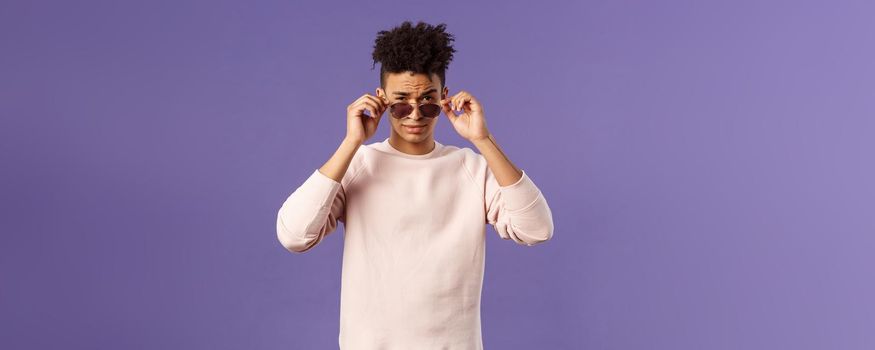 Portrait of sassy and confident young judgemental man, taking-off glasses squinting suspicious, security checking out and judging person near club door, standing purple background.