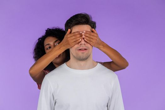 African american woman closes eyes of her beloved boyfriend before surprise him. interracial couple on purple studio background. Love, holiday, happiness concept. High quality photo