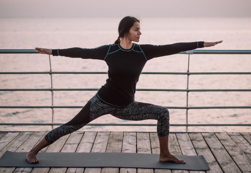 Young brunette woman doing yoga exercises on wooden sea embankment in the morning. Girl in black sports costume. Stretching, practice, healthy lifestyle concept.