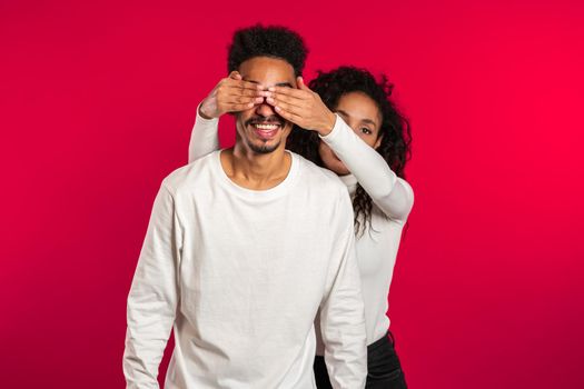 African woman closes eyes of her beloved boyfriend before surprise him. Couple in white on red studio background. Love, holiday, happiness concept