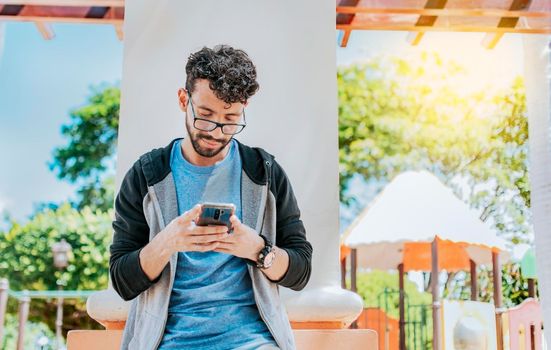 Handsome guy in glasses leaning on a wall chatting on his cell phone, Teenage male leaning on a wall sending a text message, Smiling handsome man using a phone leaning on a wall in a park