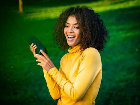 Modern trendy african american girl listening to music by wireless portable speaker.Young beautiful woman enjoying, dancing in park