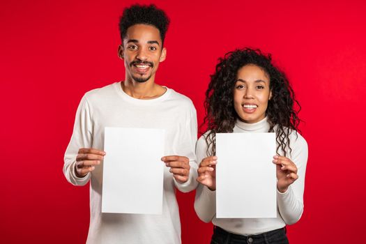 Portrait of young handsome african american man and woman couple holding white empty paper blanks on red studio background. Copy space. Match with separate sign.