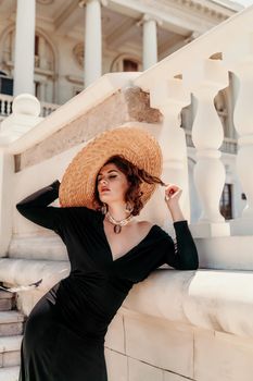 Fashion photo of a beautiful model in an elegant hat and black dress posing against the backdrop of a building with columns