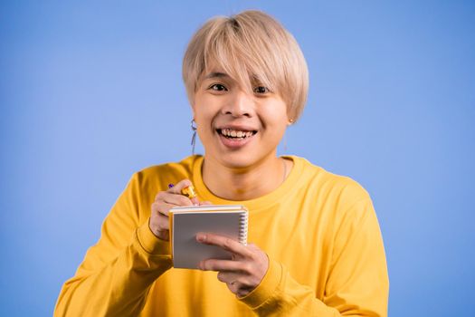 Asian man making notes in planner, guy holding pen. He writes future plans and to-do list in notebook for week or month. Keeping personal diary on blue studio background. High quality