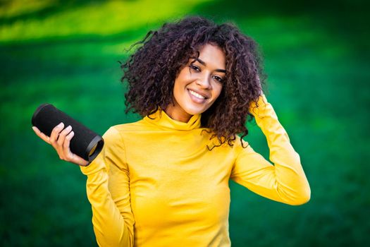 Modern trendy african american girl listening to music by wireless portable speaker.Young beautiful woman enjoying, dancing in park