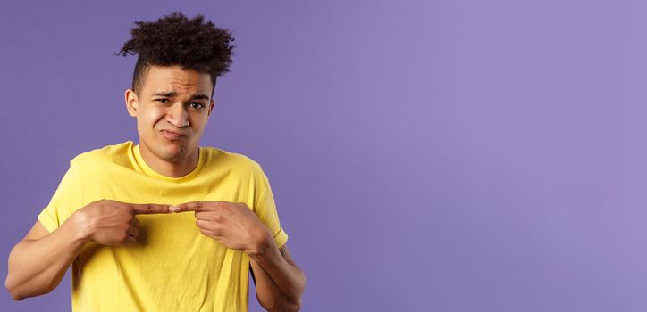 Close-up portrait of shy and modest young silly hispanic man trying say something but being too insecure, grimacing and frowning, look timid, two fingers touching pose, purple background.