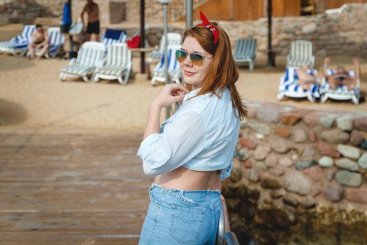 attractive smiling young adult woman in sunglasses stands with her back on the beach of the mediterranean sea in Turkey.