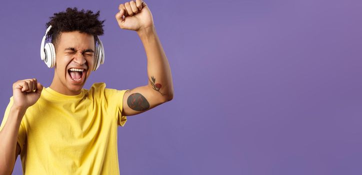 Close-up portrait of cheerful, happy young dancing guy lift hand up singing along, close eyes and smiling upbeat as listening awesome song in headphones, enjoying music, purple background.