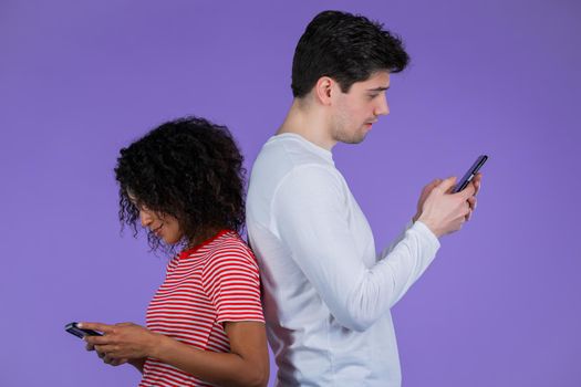 Interracial couple stand with their backs to each other and print messages or surf Internet from mobile phones. Technology, distancing, smartphone, social networks concept.