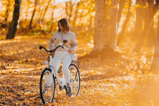 Beautiful portrait of young woman cycling alone in autumn park. Sunny day, golden leaves in forest. Trendy lady on vintage white bicycle, healthy lifestyle, aesthetic scene. High quality photo