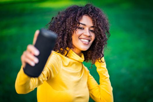 Modern trendy african american girl listening to music by wireless portable speaker.Young beautiful woman enjoying, dancing in park