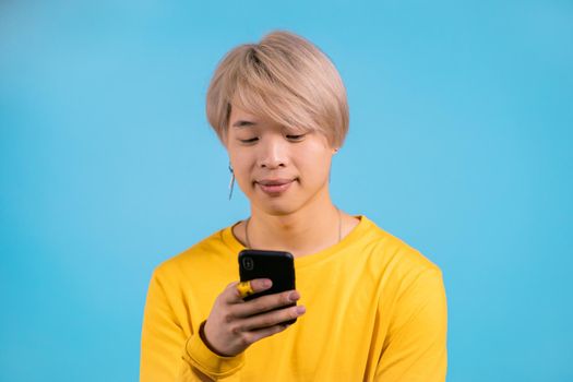 Handsome korean man using smartphone on blue studio wall. Blond guy in yellow outfit using modern technology - apps, social networks