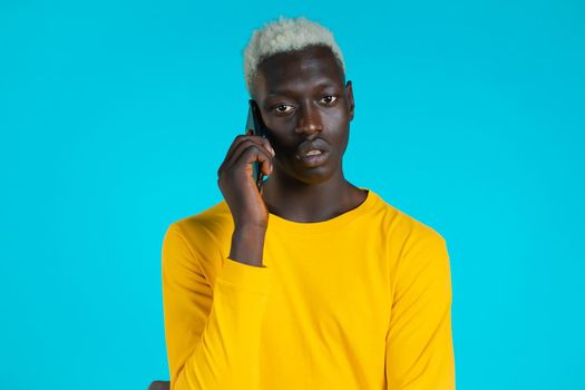 Young afro man speaks serious on phone. Guy holding and using smart phone. Blue studio background