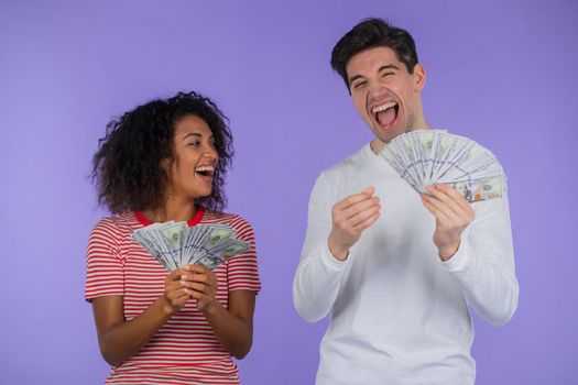 Amazed happy excited interracial couple showing money - USD currency dollars banknotes on purple wall. Symbol of success, gain, victory. African woman and white man win lottery. High quality photo