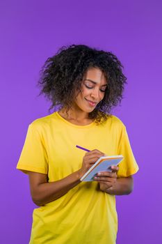 Young woman making notes in planner, african lady holding pen. She writes future plans and to-do list in notebook for week or month. Keeping personal diary on purple studio background. quality photo