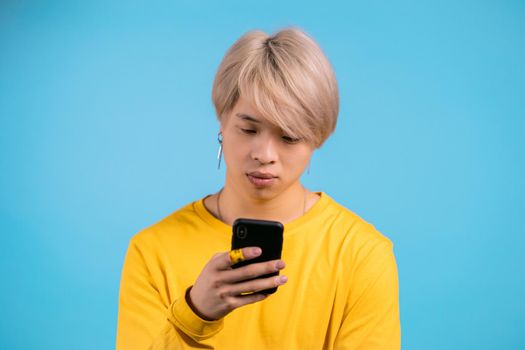 Handsome korean man using smartphone on blue studio wall. Blond guy in yellow outfit using modern technology - apps, social networks