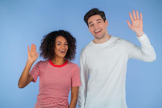 Interracial friendly couple - white man and african woman waving hands - goodbye, chao, adios. Parting, say bye to camera on blue studio background. High quality
